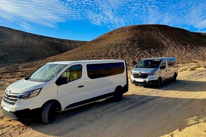Two white vans parked on a dirt road in a desert landscape with hills in the background. 3