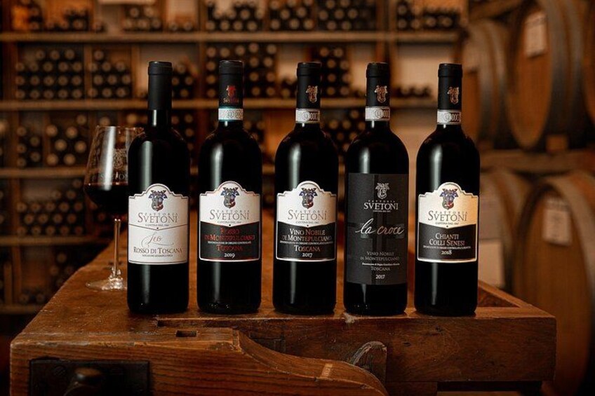 Five bottles of wine on a wooden table with a wine rack in the background. 3