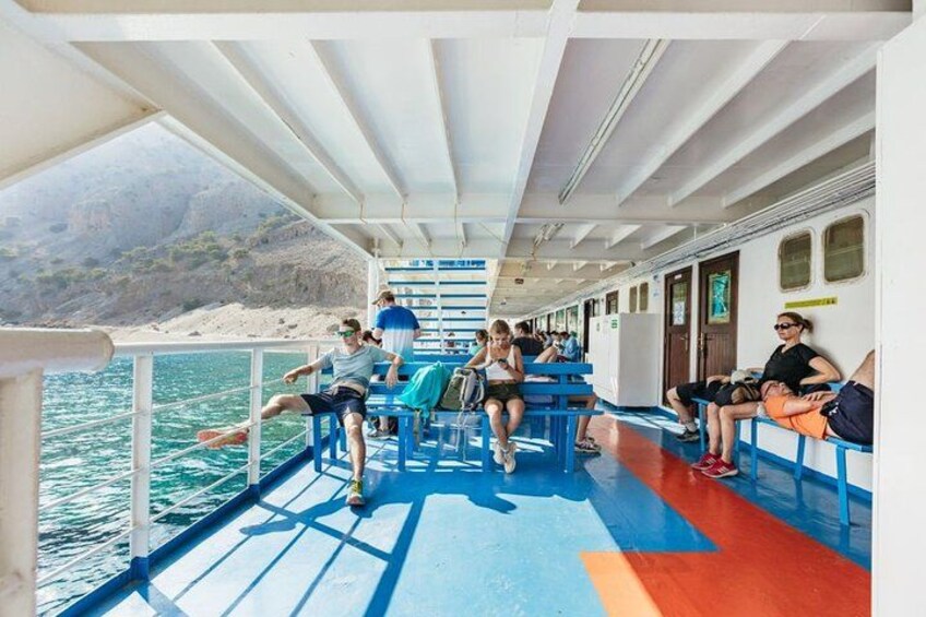 A ferry with passengers seated on blue benches, a railing, and a view of the sea and a beach. 2