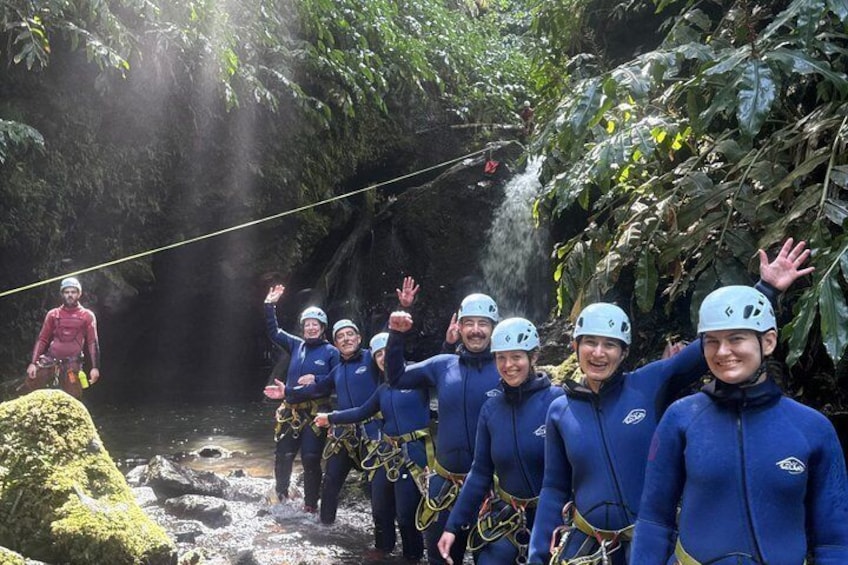 Een groep mensen in blauwe neopreenpakken en helmen bereidt zich voor op een wateractiviteit, met een touwbrug en een waterval op de achtergrond. 5