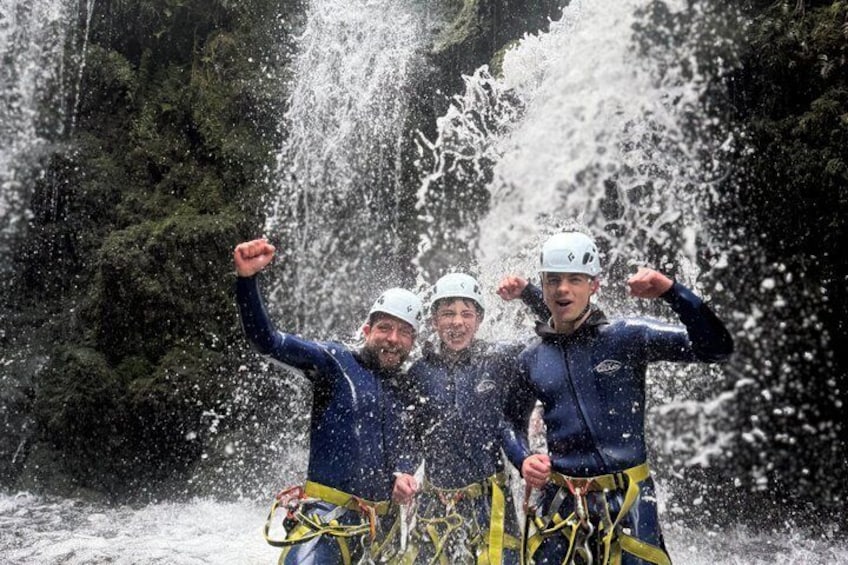 Drie mensen in blauwe neopreenpakken en helmen vieren, met een waterval op de achtergrond. 2