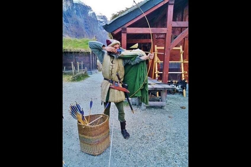 Eine Person in mittelalterlicher Kleidung mit Bogen und Pfeilen, stehend neben einem Pfeilkorb. 3