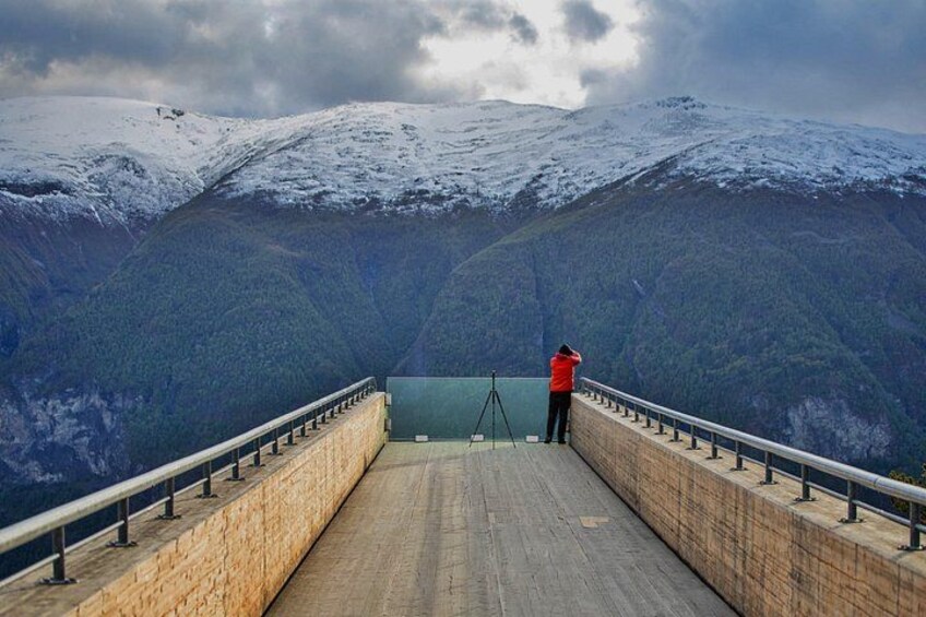 Eine Person in einer roten Jacke macht ein Foto mit einem Stativ auf einer hölzernen Plattform mit Geländern, mit Blick auf verschneite Berge. 5