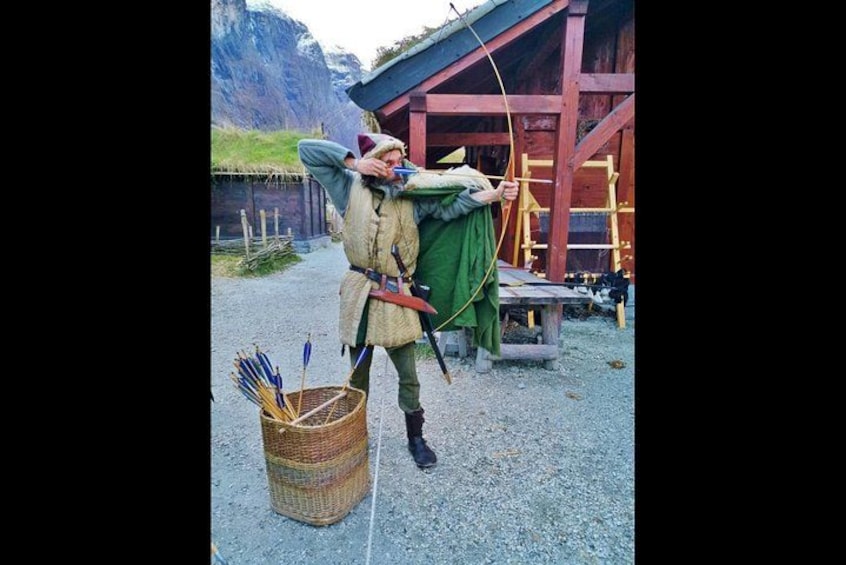 Una persona in costume medievale con arco e frecce, in piedi di fronte a una struttura in legno. 2