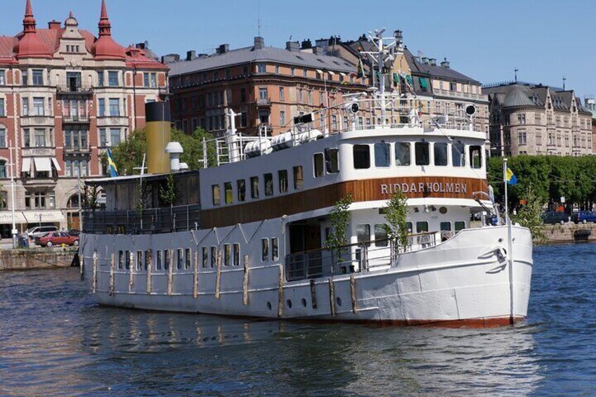 En stor vit båt som heter Riddarholmen ligger i en flod, med byggnader i bakgrunden. 1