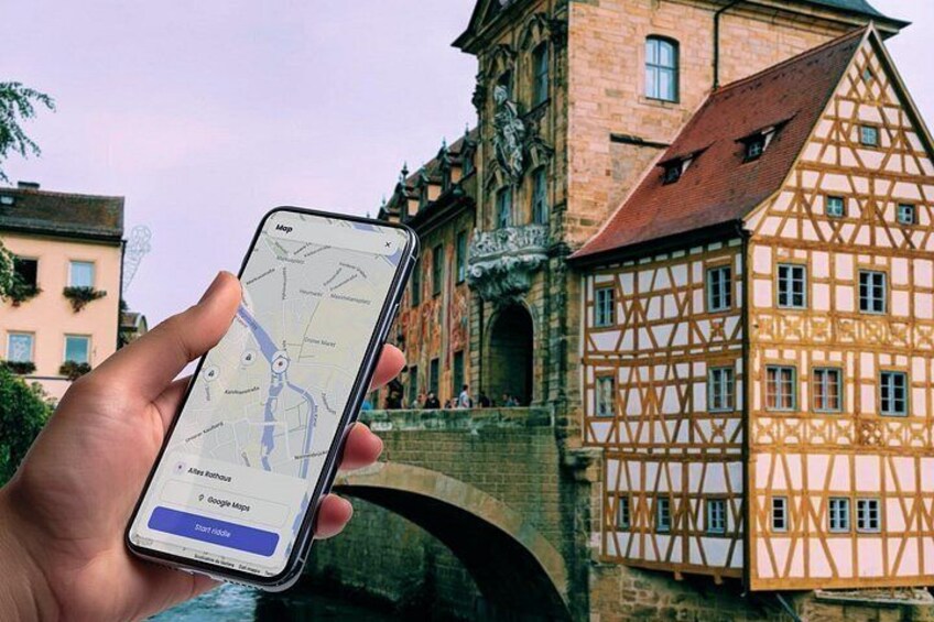Une main tenant un smartphone sur lequel est ouverte une application de cartographie, affichant un lieu avec un pont et, en arrière-plan, des bâtiments traditionnels à colombages. 1
