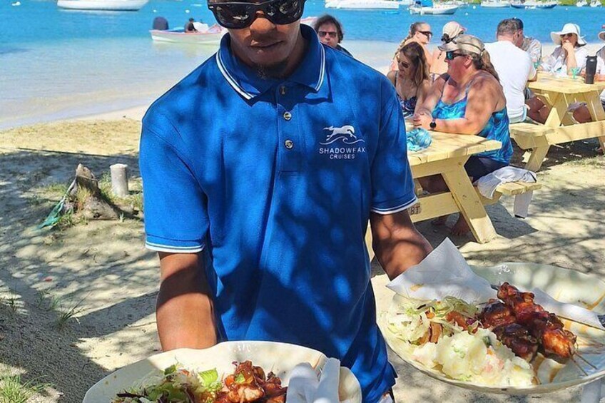 A beach scene with people dining at wooden tables, a man in a blue shirt serving food, and boats in the water. 3