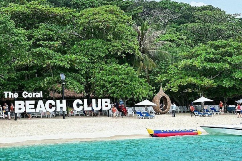 Ein Strand mit einem Schild, auf dem 'The Coral Beach Club' steht, Palmen und Liegestühlen. 5