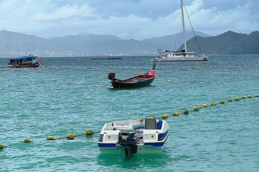 Ein ruhiges Meer mit mehreren Booten, darunter ein Segelboot und ein Motorboot, vor einer Küste mit Bergen im Hintergrund. 4