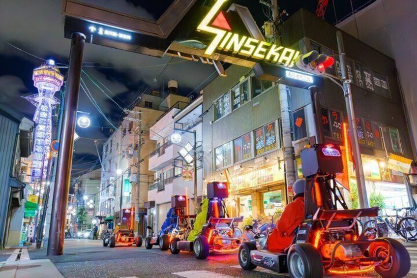 A brightly lit urban street with neon signs, a Ferris wheel, and colorful go-karts. 1