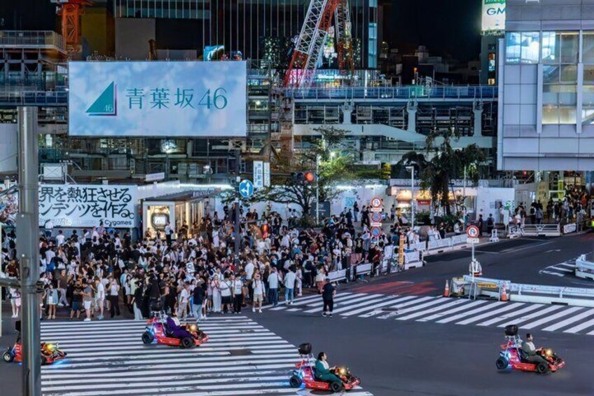 A crowded street scene with a large crowd gathered at a crosswalk, a sign for '青葉坂46', and a building with a visible 'GM' sign. 2