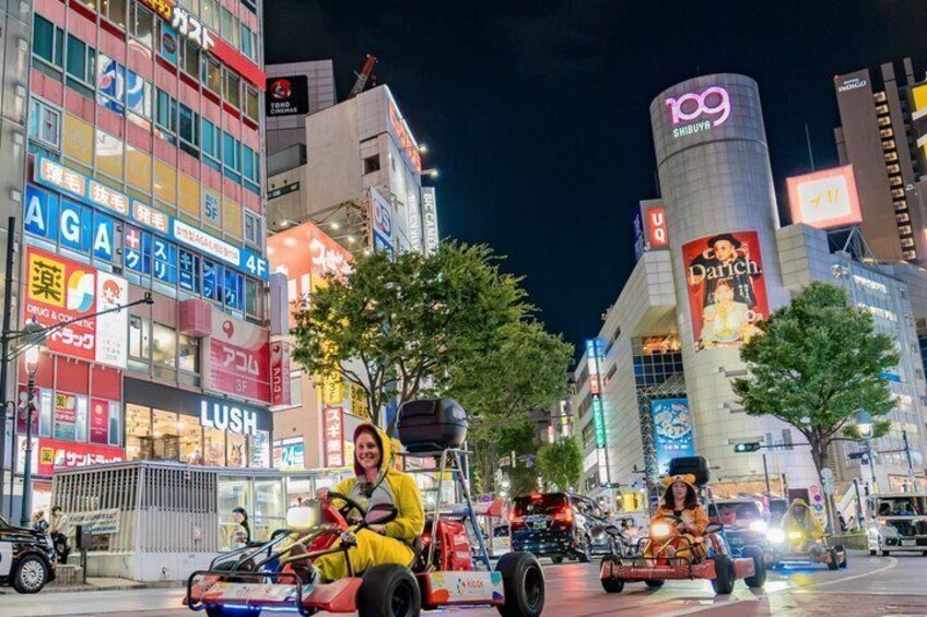 A nighttime urban street with go-karts, illuminated billboards, and a prominent 109 Shibuya building. 1