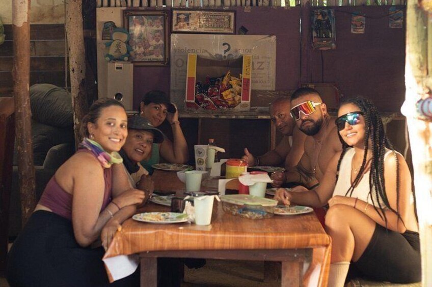 A group of people sitting around a wooden table with various items on it, including plates, cups, and a box. 3