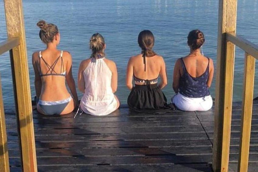 Four women sitting on a wooden dock by a calm body of water. 3