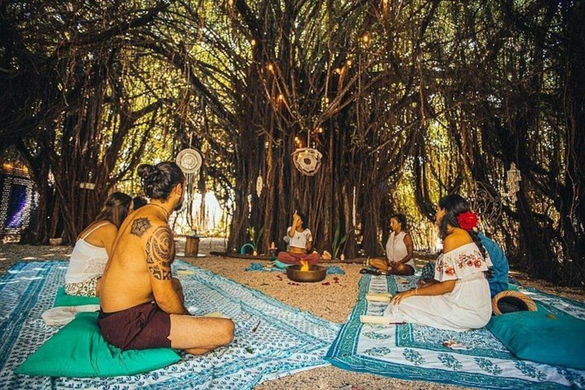 A group of people sitting on mats under a tree, engaged in a circle. 1