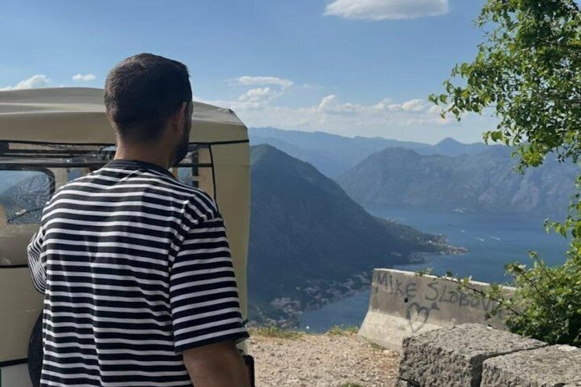 A person standing by a vehicle overlooking a mountainous landscape with a lake and a sign with graffiti. 4