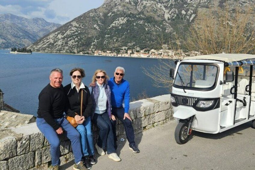 Four people sitting by a lake with mountains in the background and a white vehicle parked nearby. 2