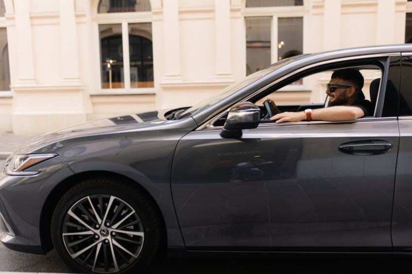 A person driving a dark-colored car with a visible steering wheel and side mirror. 2