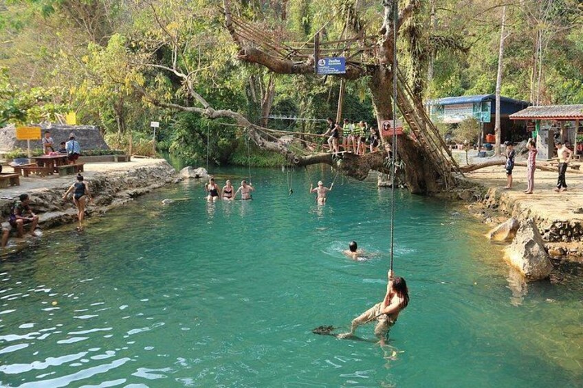 Ein natürliches Schwimmrevier mit Personen im Wasser, ein Baum, der über das Wasser ragt, und eine Holzbrücke. 1