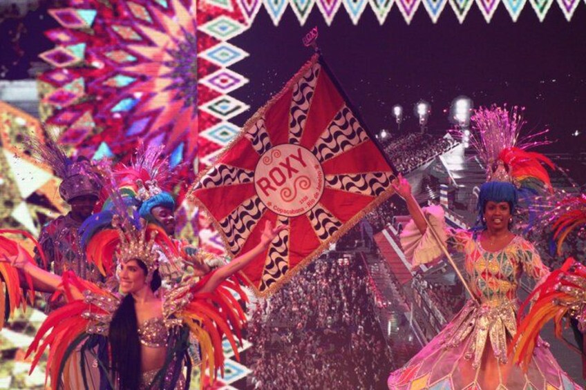 A person holding a flag with a circular emblem, surrounded by colorful decorations and a crowd. 4