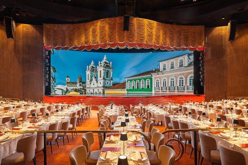 Un salón de banquetes con mesas dispuestas para un evento formal, una pantalla grande mostrando un paisaje urbano y un escenario con cortina dorada. 2