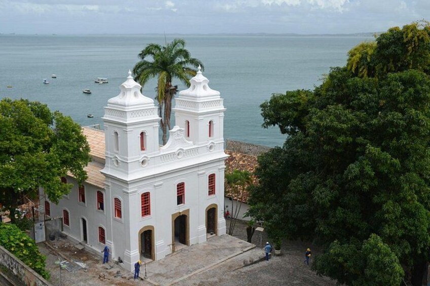 A white colonial-style building with red accents, palm trees, and a view of the sea with boats. 1