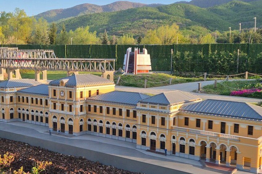 A large building with multiple arched windows and a clock tower, surrounded by greenery and hills. 2