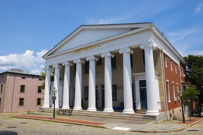A neoclassical building with white columns and a pediment, surrounded by a cobblestone street and other historic buildings. 4