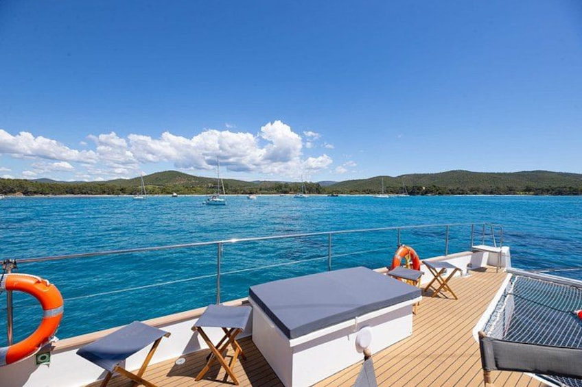 A boat deck with lounge chairs, a lifebuoy, and a view of a calm sea with boats and hills in the background. 4