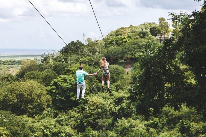 Una persona suspendida en el aire por un tirolina, otra persona sosteniendo la cuerda, y un paisaje verde exuberante con árboles y una vista distante del océano. 4