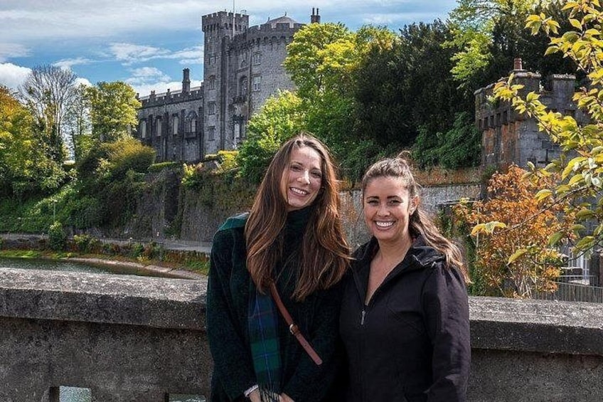 Dos personas de pie frente a un castillo histórico con muro de piedra y árboles al fondo. 5