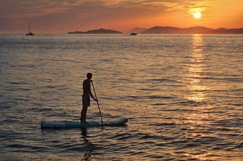 A person paddleboarding on calm water during sunset with distant boats and a hilly landscape. 1