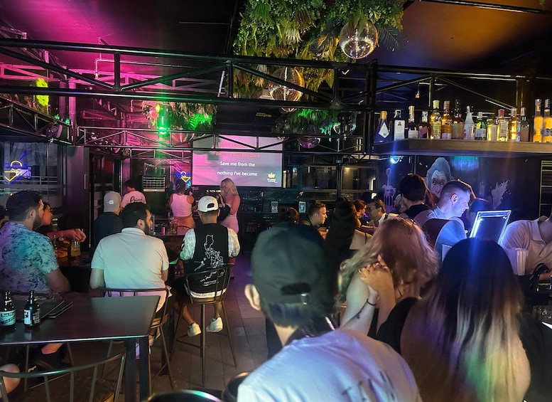 Un bar affollato con persone sedute e in piedi, un palco per DJ e un bancone bar pieno di bottiglie. 2
