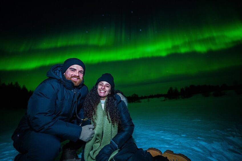 Two people sitting on snow with the aurora borealis in the background. 2