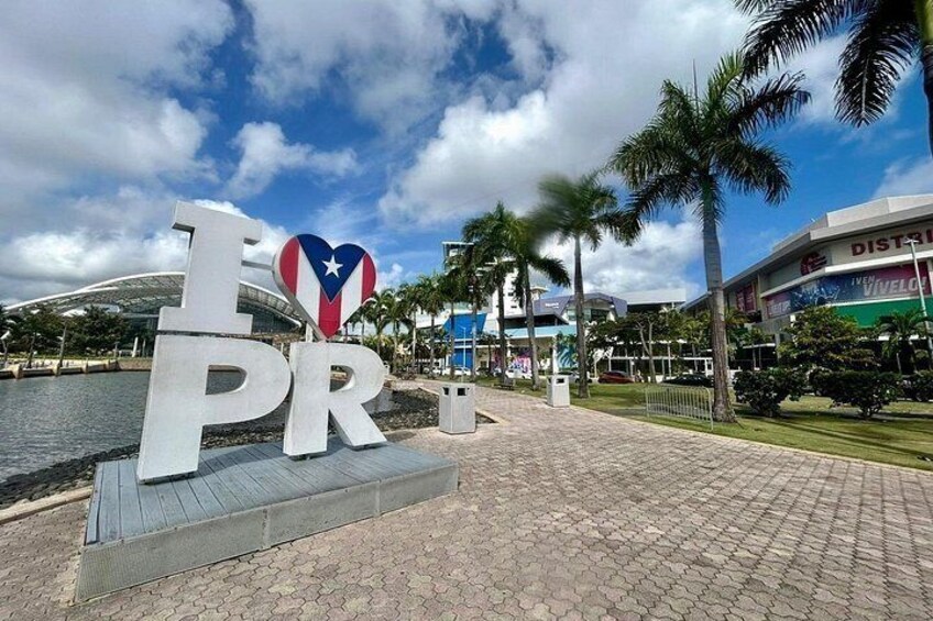 A sign with 'I ♥ PR' and a heart-shaped Puerto Rico flag, palm trees, a paved walkway, and a building with 'DISTAL' signage. 2