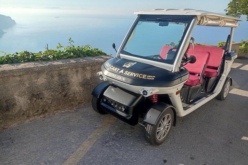 A golf cart with 'AMALFI COAST SERVICE' written on the front, parked on a road with a scenic view of the sea in the background. 2