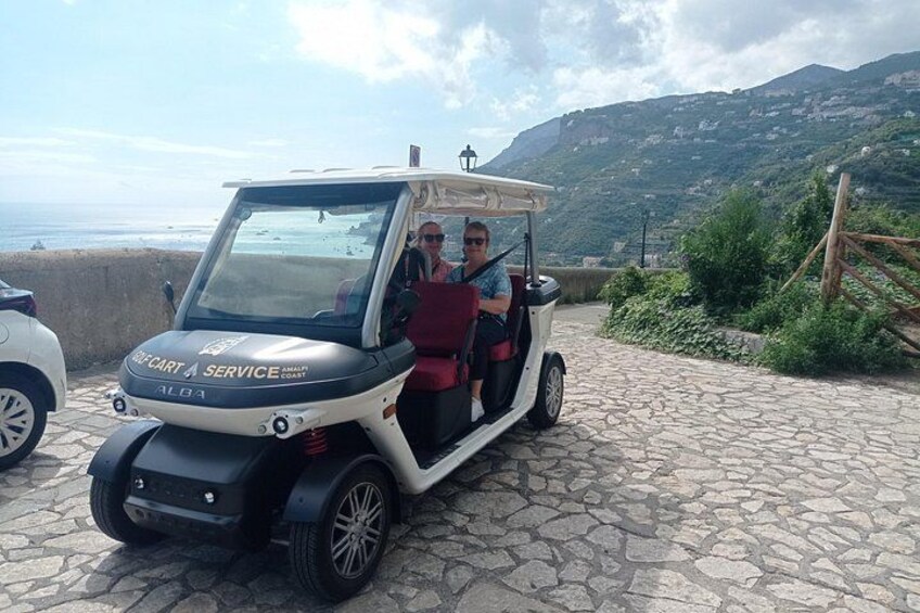 A golf cart with two passengers, parked on a stone path with a coastal view. 3