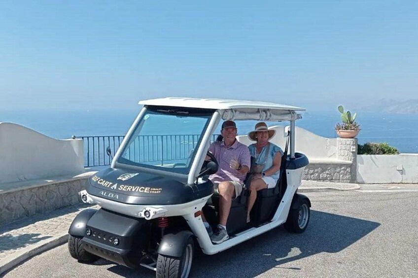 A golf cart with 'ALGA SERVICE' on the front, two people sitting inside, and a coastal landscape with a clear sky in the background. 1
