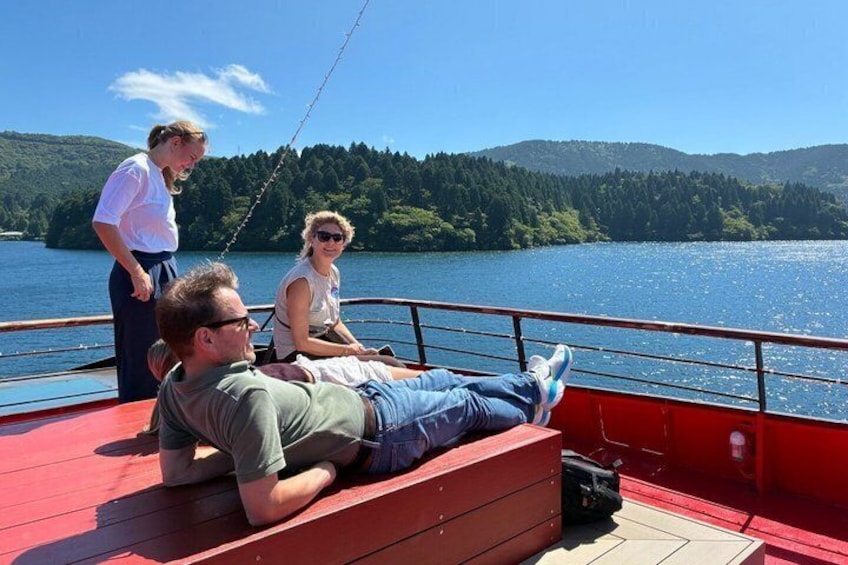 A person lying on a red deck, another person standing, and a third person sitting, all on a boat with a lake and forested hills in the background. 4