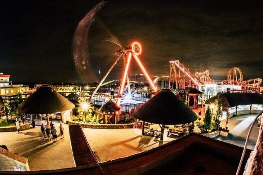 A nighttime amusement park with a Ferris wheel, roller coasters, and thatched-roof structures. 2