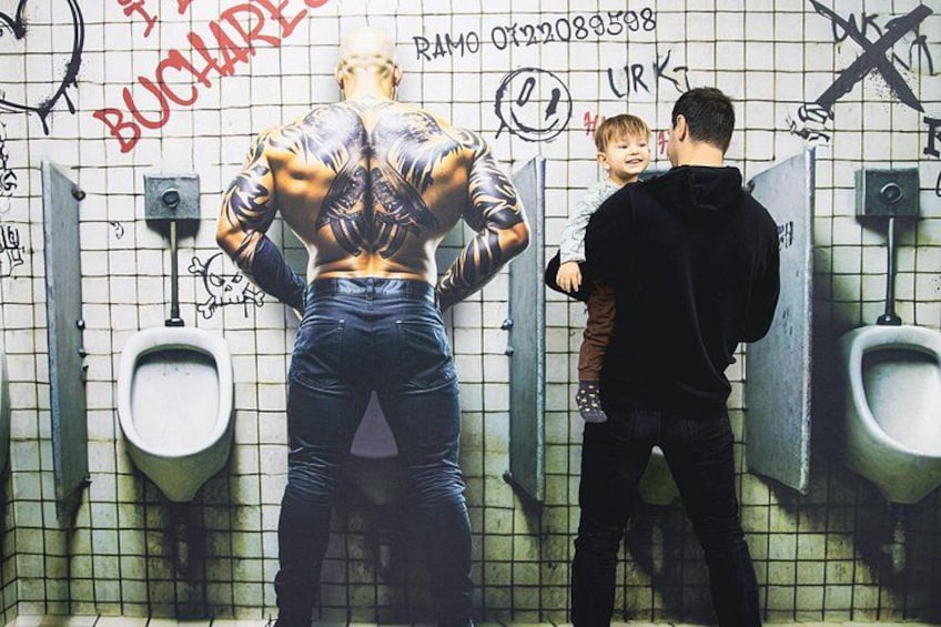 Un homme tatoué se tient dans un urinoir, les murs étant couverts de graffitis. 3