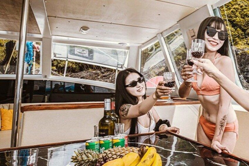 Dos personas brindando con bebidas en un barco, una mesa con botellas y un plátano, y vistas del agua y árboles. 3