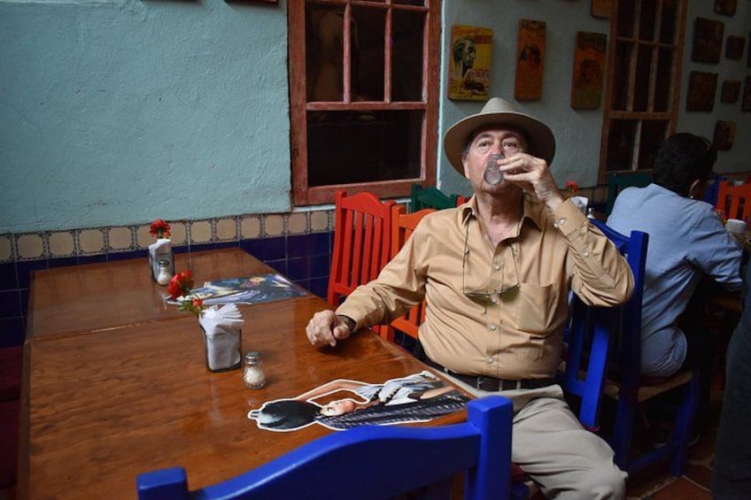 A man sitting at a wooden table in a restaurant with blue chairs, a window, and posters on the wall. 3