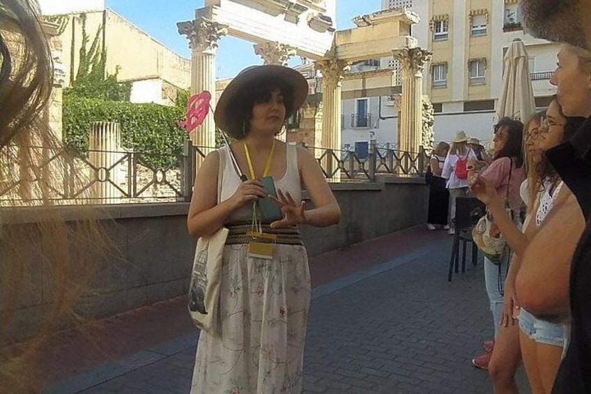 A woman in a white dress and hat, holding a phone, standing on a paved walkway with a railing and buildings in the background. 3