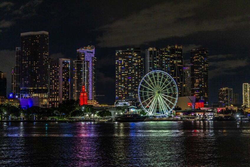 A city skyline at night with illuminated buildings and a large Ferris wheel reflecting on the water. 1
