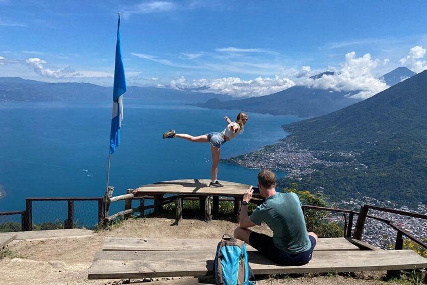 A person performing a balancing act on a wooden platform with a flag, overlooking a mountainous landscape and a body of water. 1