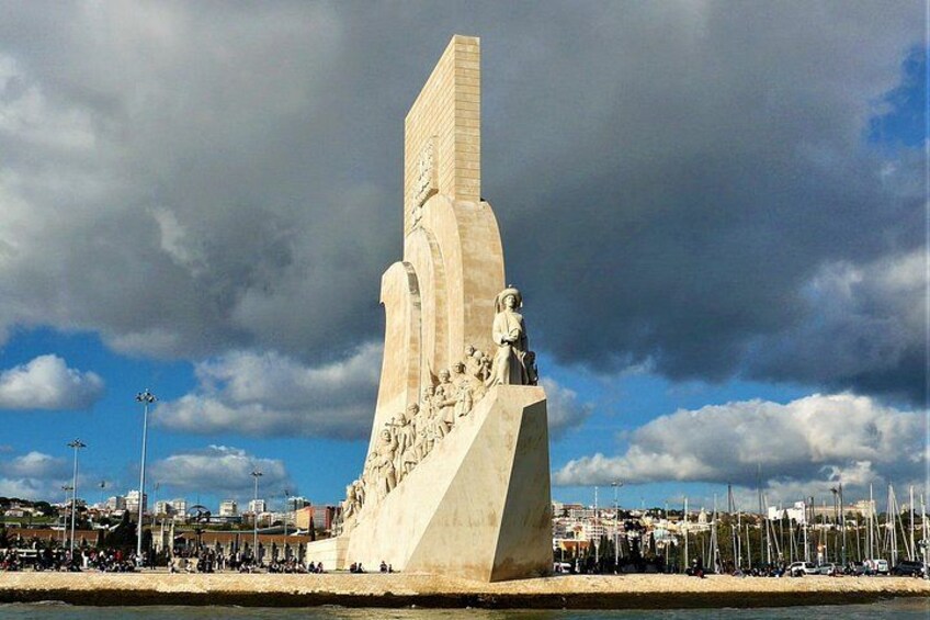 Ett stort monument med en triangulär bas och en hög, smal struktur ovanpå, omgivet av en strandpromenad och segelbåtar. 3