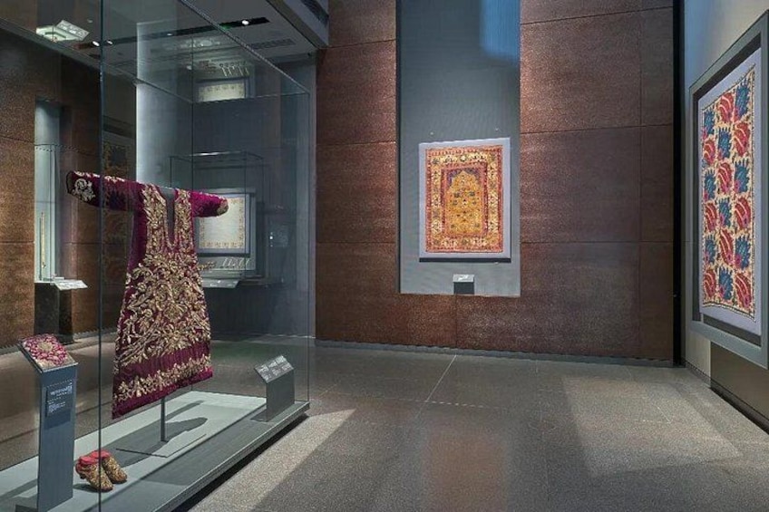 Ein Museuminneres mit einer Glastreppe, die ein traditionelles Kleidungsstück, einen gerahmten Teppich und ein gerahmtes Textilstück zeigt. 5