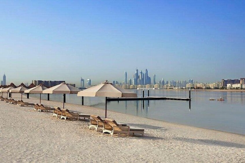 A beach with lounge chairs and umbrellas, a calm water body, and a city skyline in the background. 1