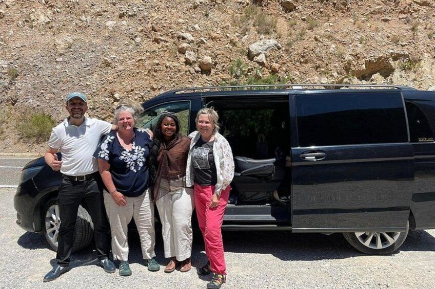 Cuatro personas frente a un SUV negro con la puerta abierta, un cerro rocoso al fondo y una carretera pavimentada. 5